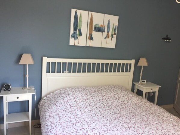 Une chambre avec un lit blanc, deux tables de chevet avec des lampes, une œuvre d’art accrochée au mur et une petite table de chevet avec une horloge.