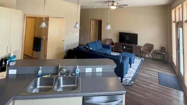 A modern kitchen with a dishwasher, a dining area with a table and chairs, a living room with a sofa and a TV, and a ceiling fan. - 2 Bed, 2 Bath Apartment at Cannery Square (Sun Prairie)