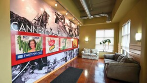 A lobby with a wall displaying vintage advertisements, a sofa, a potted plant, and a window with blinds. - 2 Bed, 2 Bath Apartment at Cannery Square (Sun Prairie)