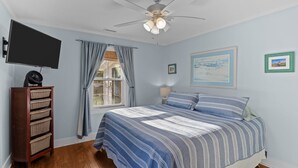 Un dormitorio con una cama, ventilador de techo, televisor, ventana con cortinas y una pared con cuadros.