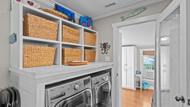 Un área de lavandería con dos lavadoras LG de carga frontal y un mueble de almacenamiento blanco en la parte superior, que contiene cestas de mimbre y objetos decorativos.