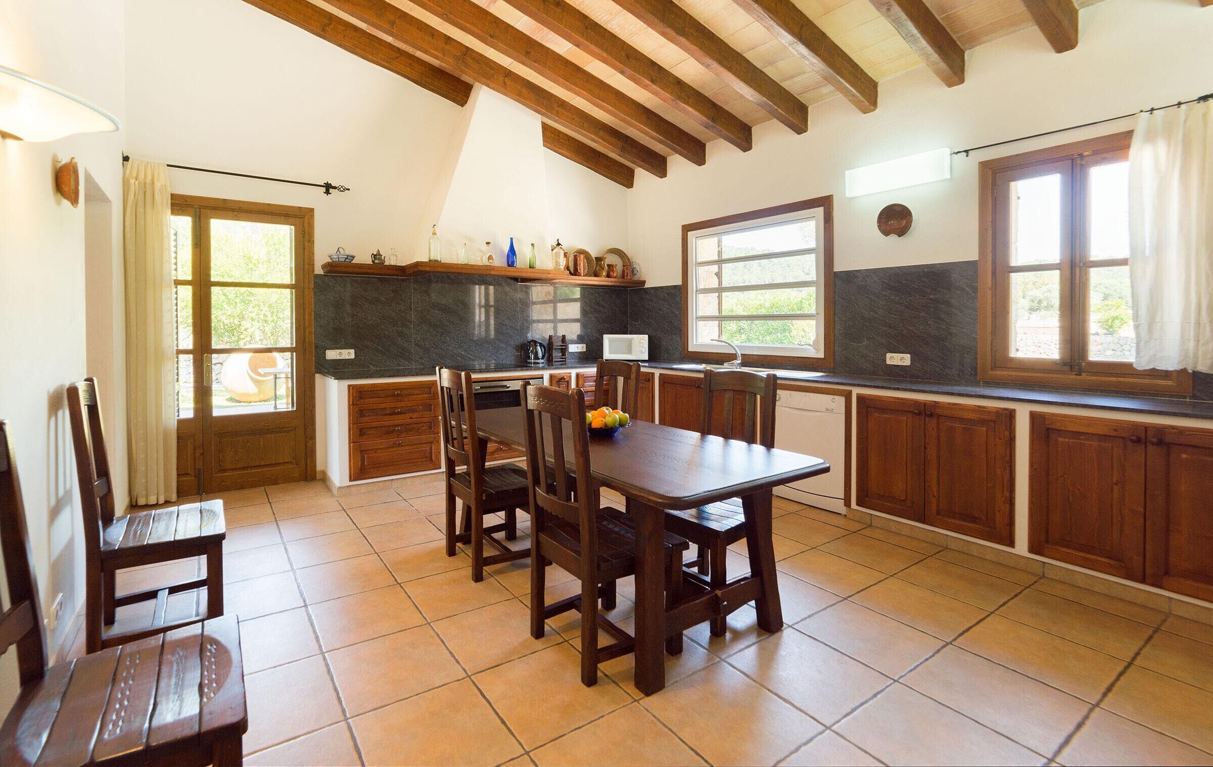 A spacious kitchen with wooden cabinets, a dining table, and chairs. There are windows providing natural light and a view of the outdoors.
