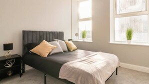 Un dormitorio con cama, mesita de noche, lámpara y dos ventanas con plantas.