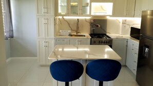 Cozinha moderna com armários brancos, bancada de mármore, fogão e dois banquetas azuis.