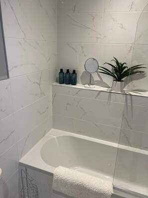 Une salle de bain avec un mur recouvert de carreaux de marbre, une baignoire blanche, une petite étagère avec des articles de toilette, une plante en pot et un miroir à main.
