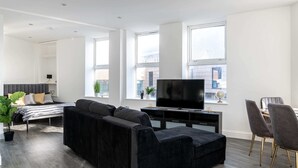 A modern living room with a black sofa, a flat-screen TV, and a dining area with chairs. - Stylish Central Studio Apartment Modern Luxury (Brighton)