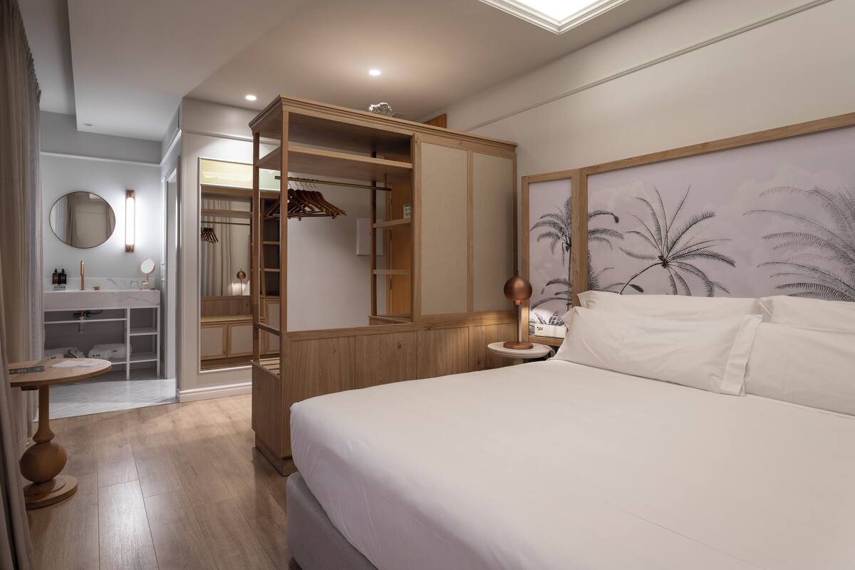 A hotel room with a bed, a round mirror, a wooden wardrobe, and a small table with a lamp.