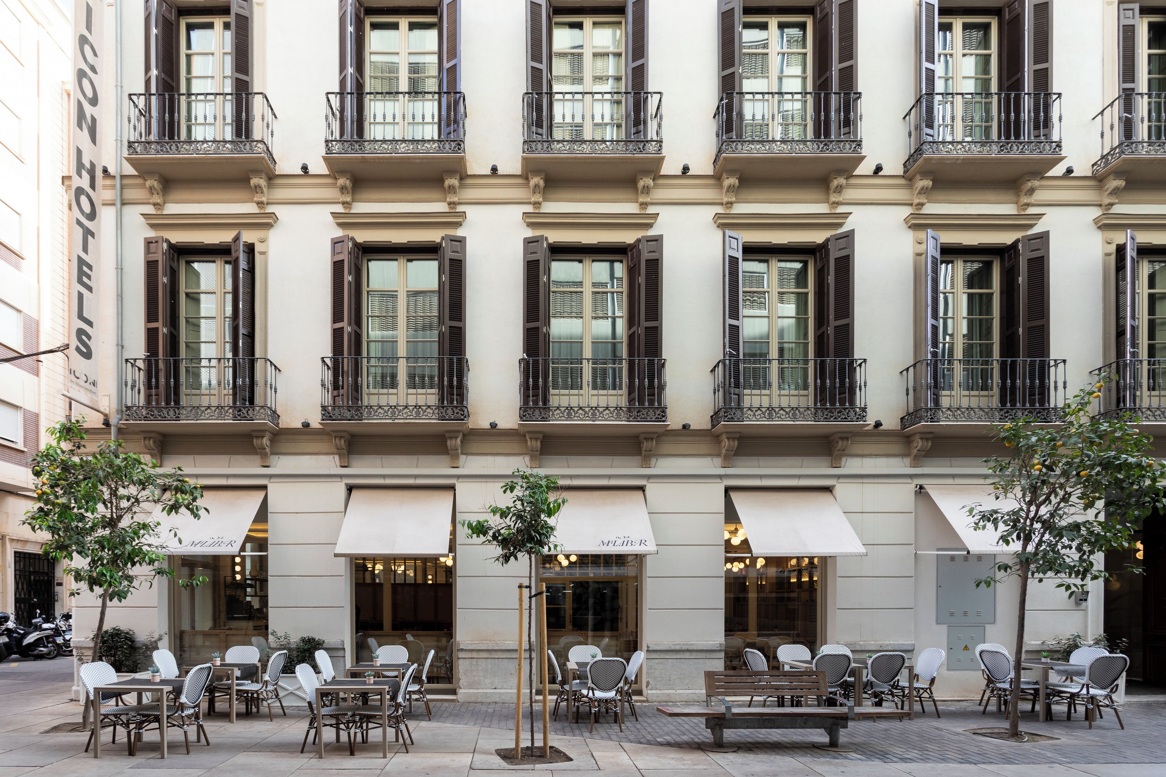 A building with multiple balconies, outdoor seating with tables and chairs, and awnings over the entrance.