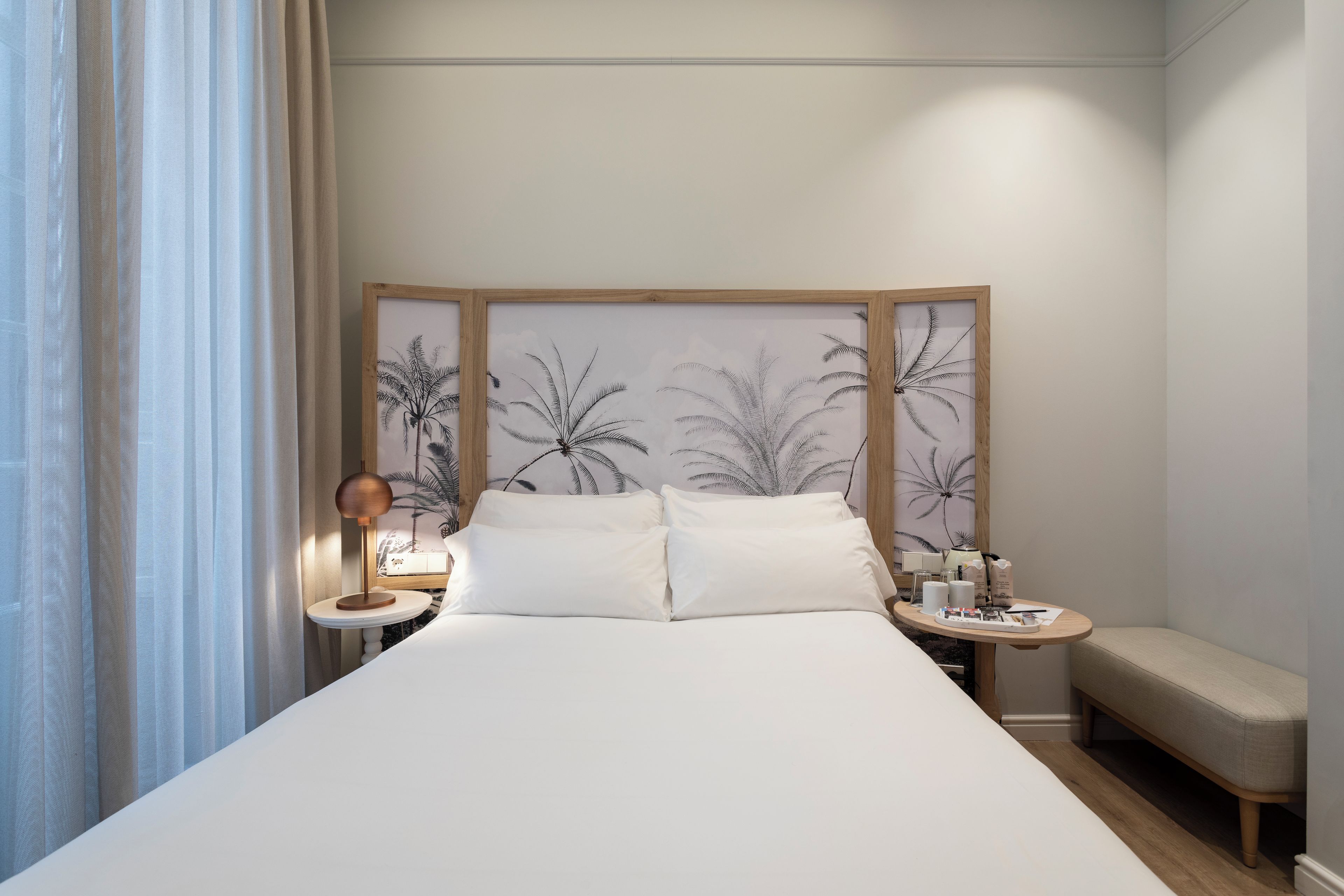 Ein ordentlich eingerichtetes Hotelzimmer mit einem großen Bett, einem Nachttisch mit Lampe, einem kleinen runden Tisch mit Tablett und einer Bank.