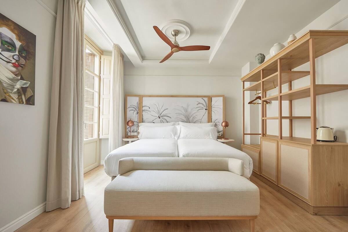 A bedroom with a bed, a wooden wardrobe, a ceiling fan, and a painting on the wall.