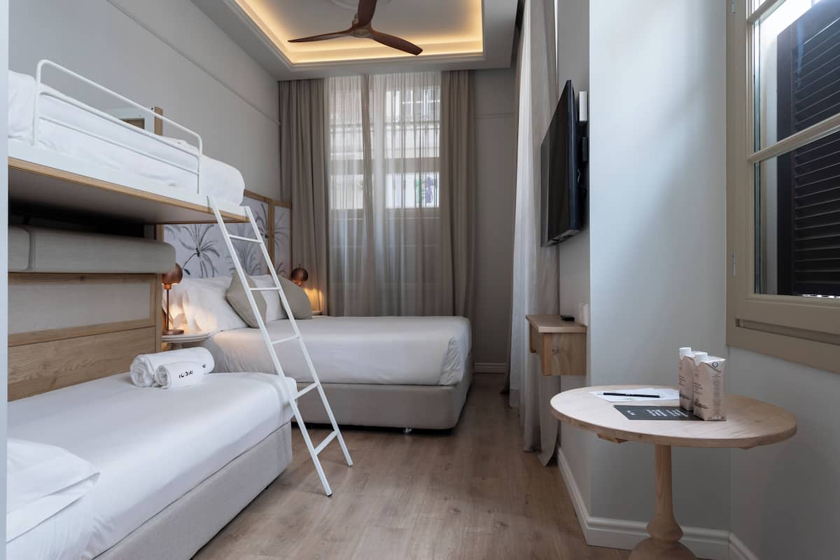 A compact hotel room with a bunk bed, a single bed, a small table, a TV, and a ceiling fan.