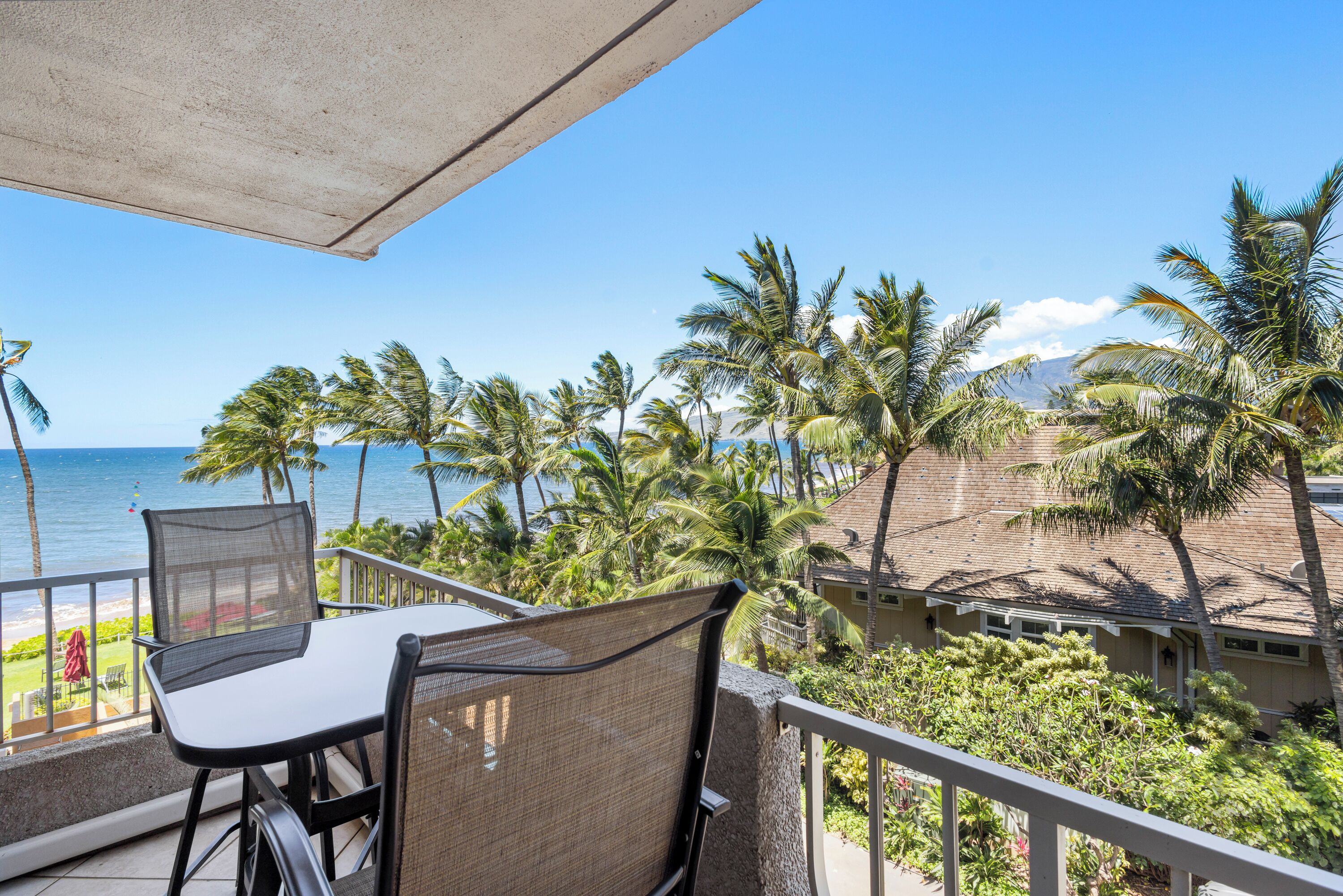 A balcony with a table and chairs, overlooking a beach with palm trees and a clear blue sky.
