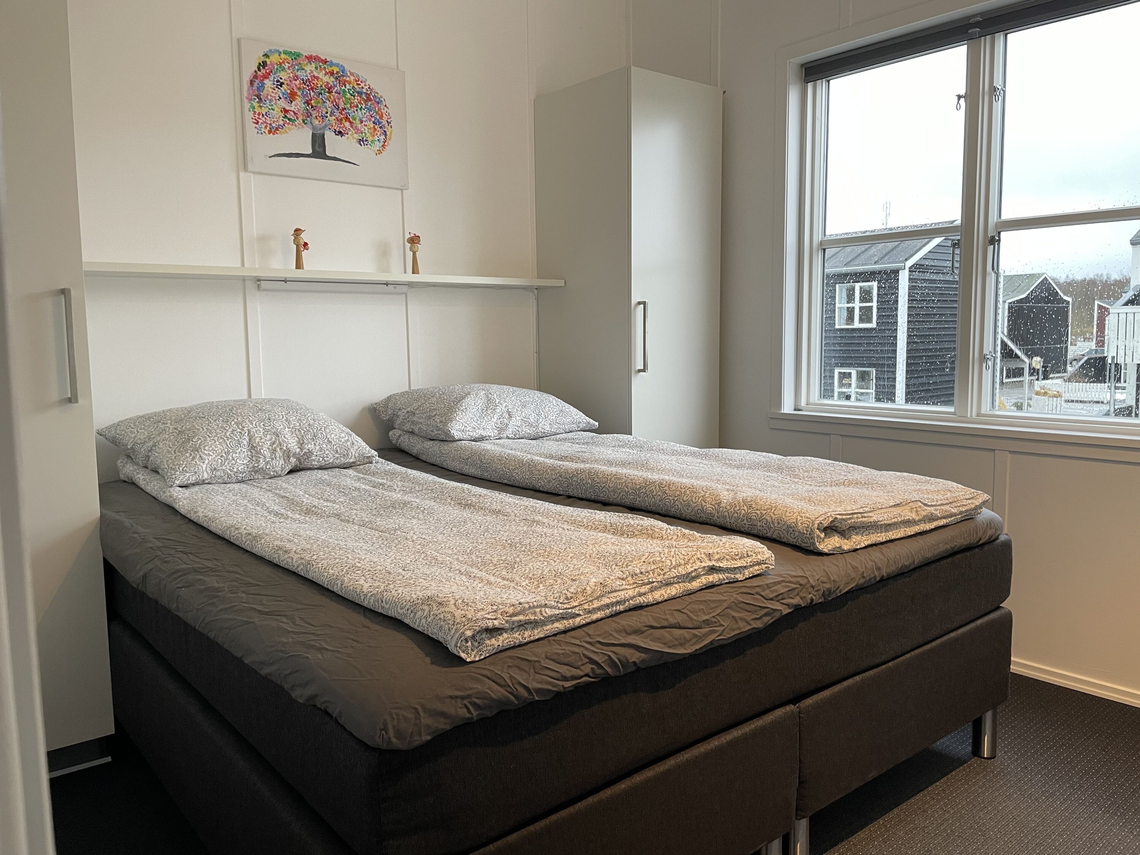 A room with two single beds, a wardrobe, and a window with a view of houses.