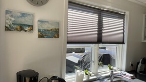 Een keuken met een koffiezetapparaat, waterkoker en een spoelbak. Twee schilderijen hangen aan de muur en er is een raam met zonwering.