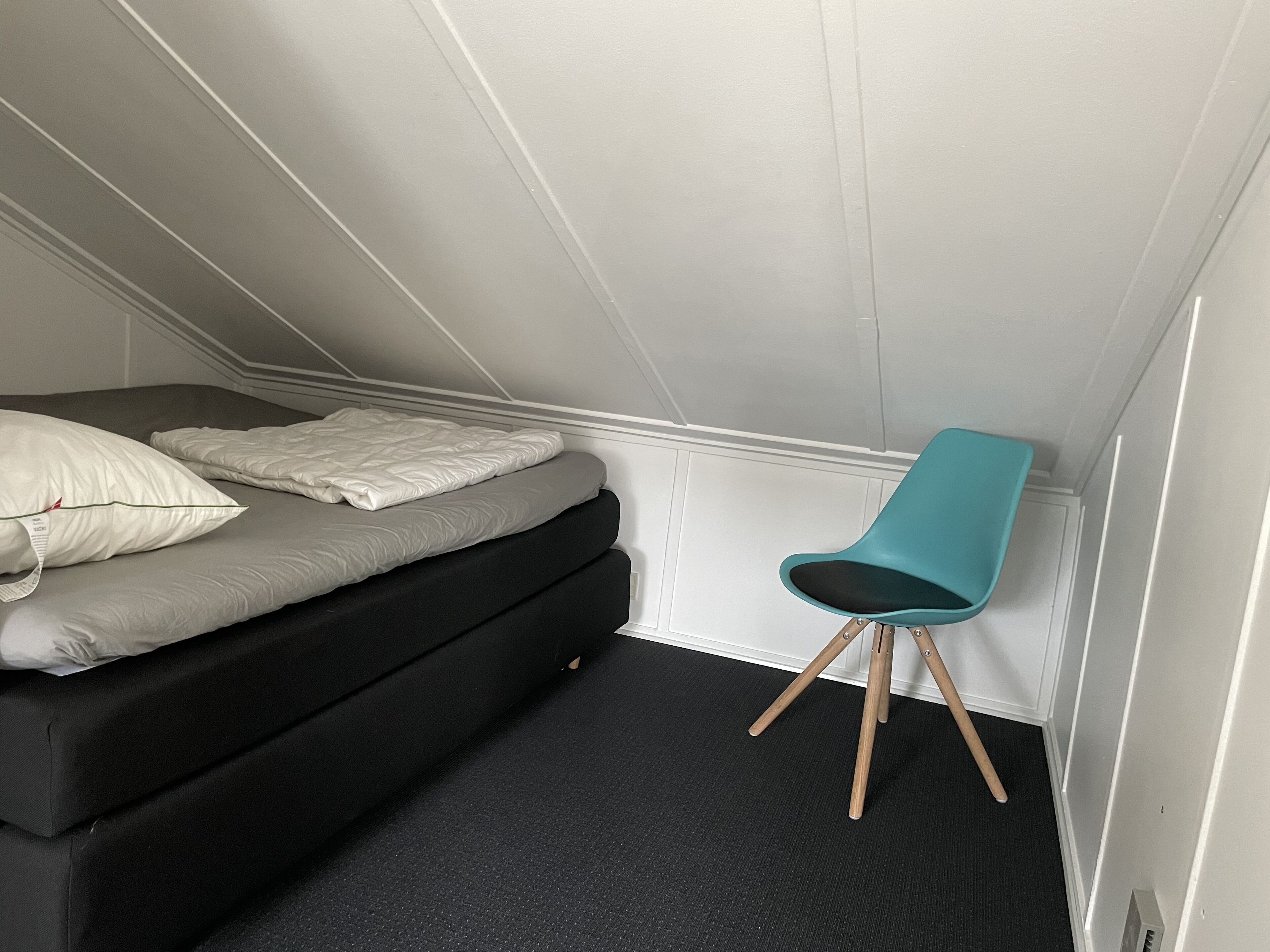 A small, compact room with a bed, a chair, and a carpeted floor.