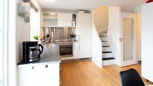 Een moderne keuken met witte kasten, een zwarte koffiezetapparaat en een roestvrijstalen oven. Er ligt een houten vloer en een witte deur leidt naar een andere kamer.