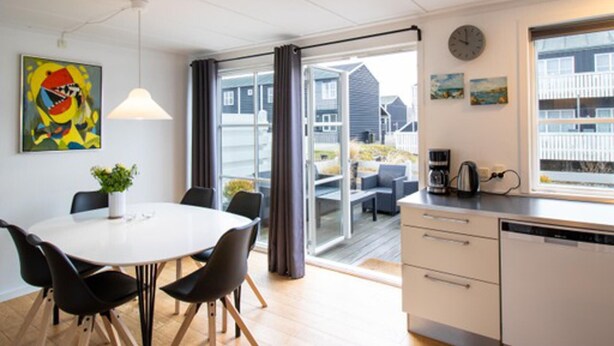 Een moderne keuken met een wit aanrecht, een koffieapparaat en een vaatwasser. Er is een eethoek met een witte tafel en zwarte stoelen. De kamer heeft een groot raam met uitzicht op huizen.