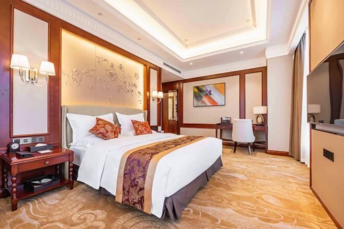 A hotel room with a large bed, a desk, and a chair. There is a nightstand with a lamp and a clock. The room has a patterned carpet and a decorative wall panel.