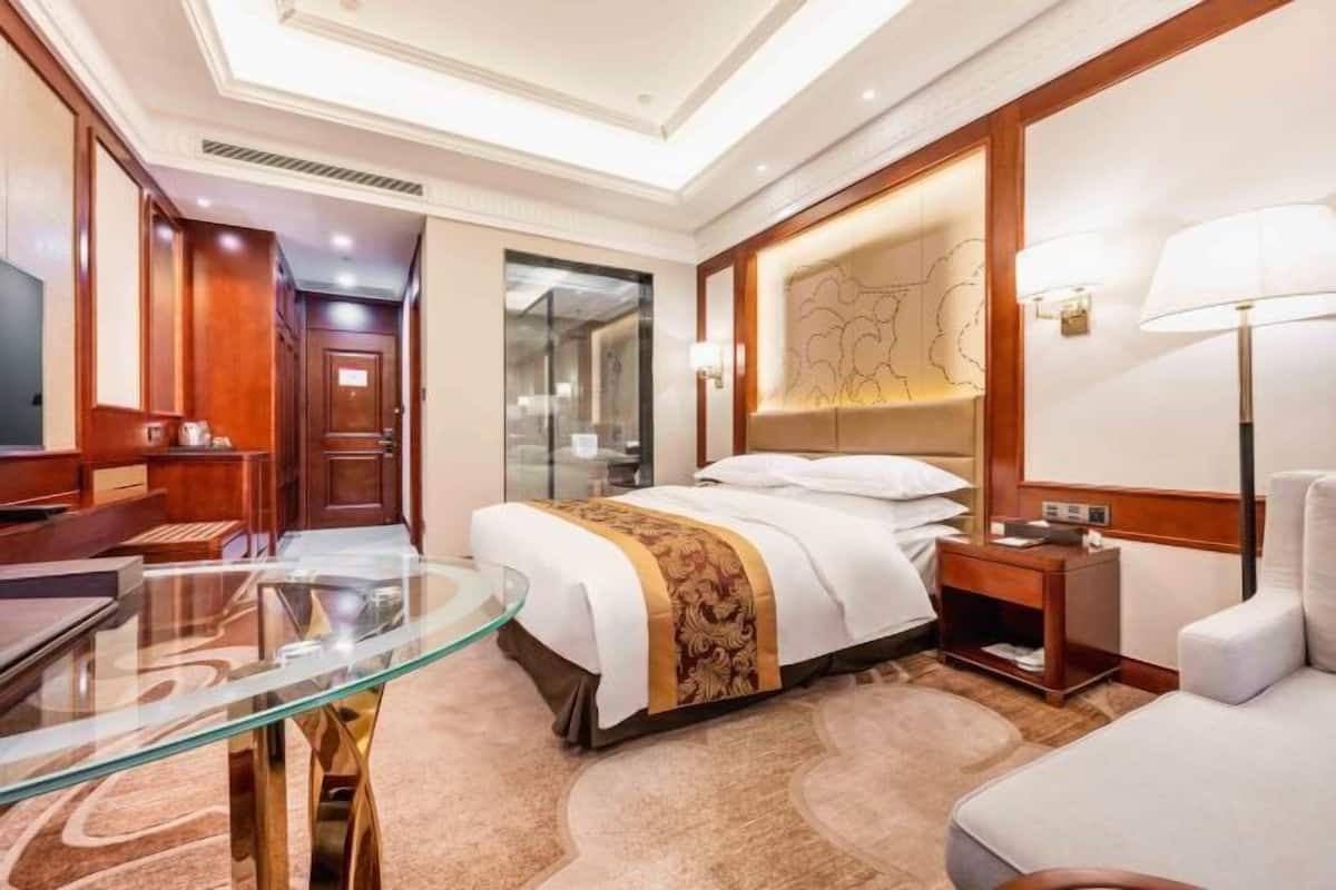 A hotel room with a bed, bedside table, lamp, and a glass table with a chair.