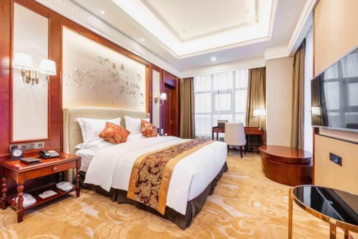 A hotel room with a large bed, bedside tables, a desk, a TV, and a view of the city.