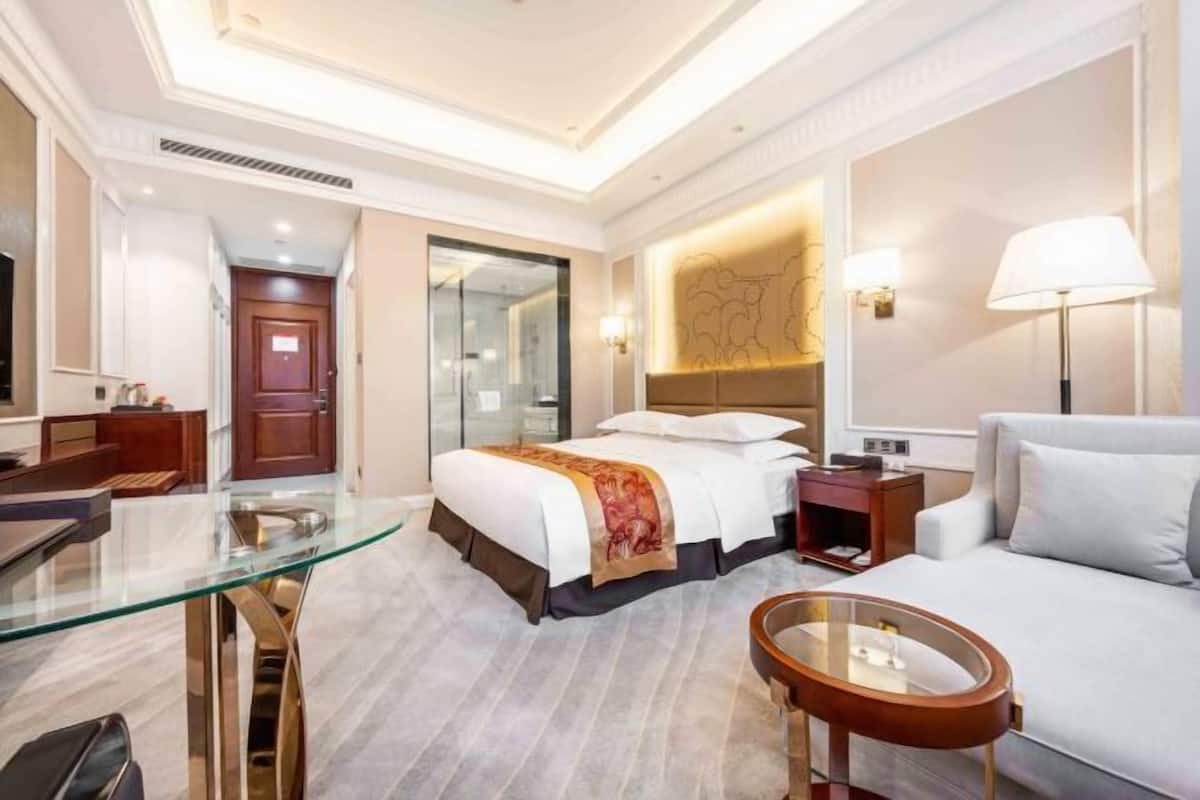 A hotel room with a glass desk, a bed with a red blanket, a sofa, a nightstand, and a lamp.