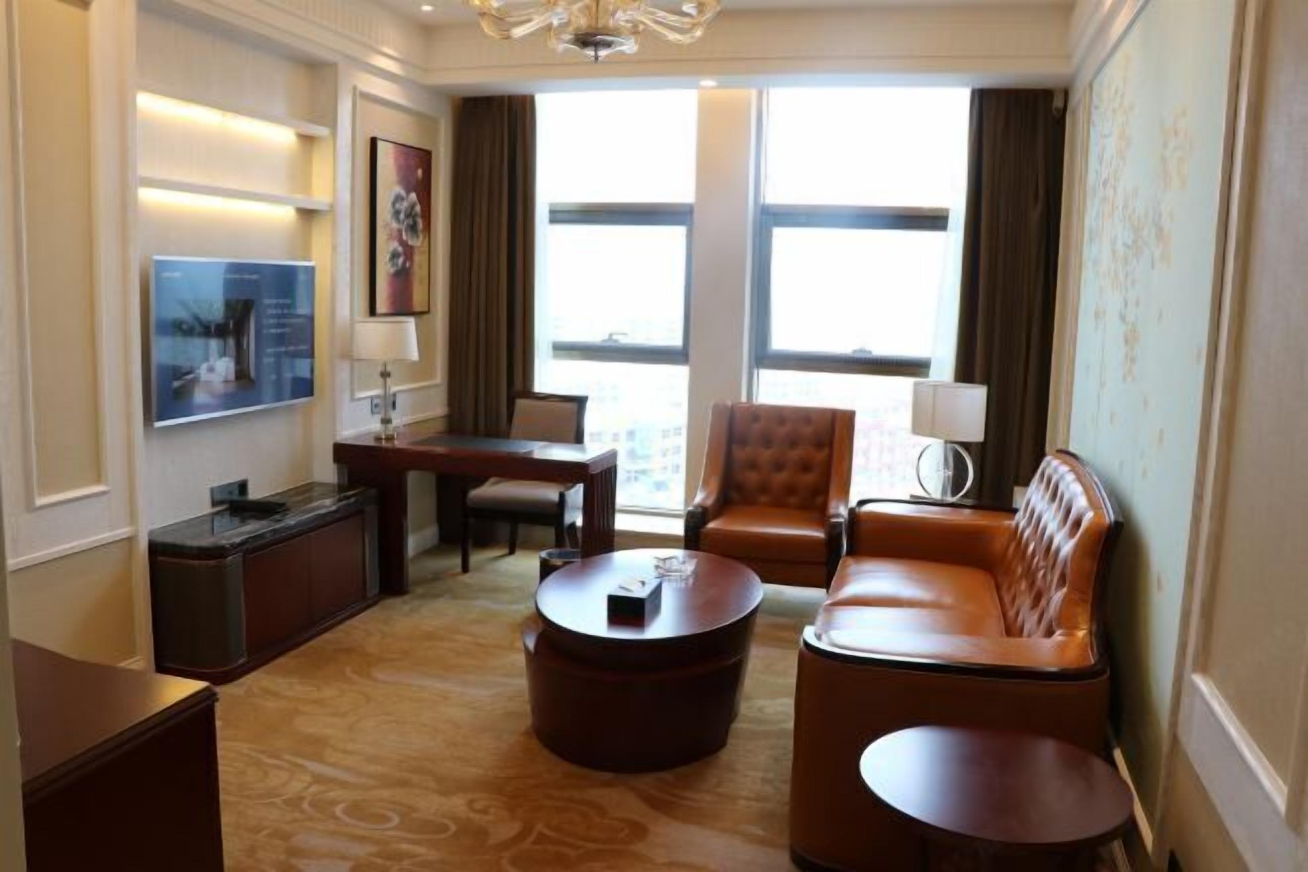 Une chambre d’hôtel avec un canapé en cuir, une table basse ronde, un bureau avec une chaise, un téléviseur à écran plat fixé au mur et une grande fenêtre avec des rideaux.