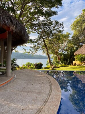 A pool area with a thatched roof structure, surrounded by lush greenery and a view of a lake with mountains in the distance. - Casa Almendra - Perfect vacation paradise (Chacala)