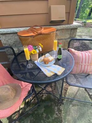 Une terrasse extérieure avec une table ronde en métal, mise avec des aliments pour le petit-déjeuner, un sac en osier et une bouteille.