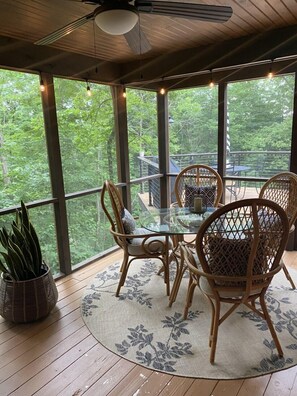 Un espace extérieur abrité, aménagé pour les repas, avec une table en verre et des chaises en osier, entouré de grandes fenêtres offrant une vue sur les arbres.