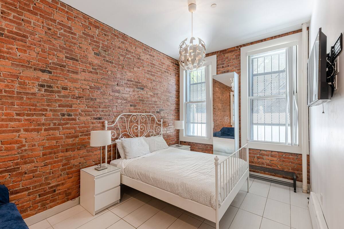 A bedroom with a brick wall, a white bed, a nightstand, a lamp, and a window with a view.