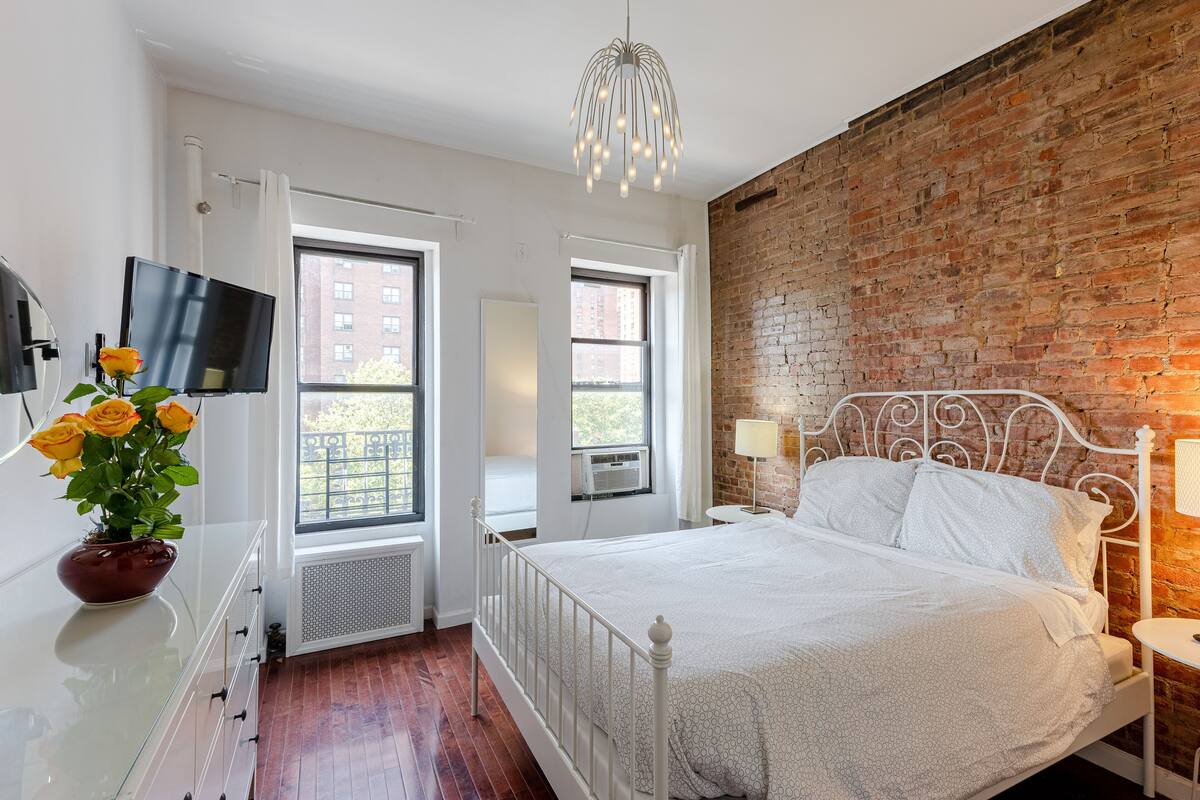 A bedroom with a brick wall, a bed with white bedding, a TV on a dresser, and a potted plant with yellow flowers.