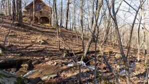 A cabin in a forested area with a wooden deck and a stone pathway leading to it. - Adventure Creek - pet friendly, hot tub, creek on site, ping pong (Boone)