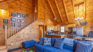 A wooden cabin interior with a blue sofa, a staircase, a kitchen area, and a dining table. - Adventure Creek - pet friendly, hot tub, creek on site, ping pong (Boone)
