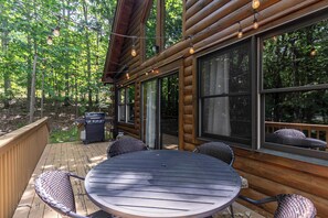 A wooden deck with a round table and chairs, surrounded by large windows offering a view of the forest.