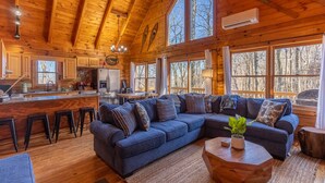A spacious wooden cabin with a large living area featuring a dark blue sectional sofa, a kitchen with wooden cabinets, and large windows offering views of the surrounding forest. - Adventure Creek - pet friendly, hot tub, creek on site, ping pong (Boone)