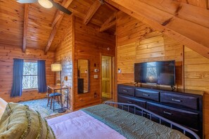 A wooden cabin bedroom with a bed, dresser, TV, desk, chair, and a window with curtains.