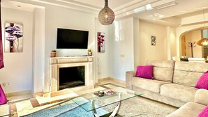 A modern living room with a flat-screen TV, a fireplace, a glass coffee table, and a beige sofa with pink cushions. - Exclusive Duplex Penthouse with panoramic views  (Marbella)