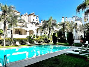 A swimming pool with lounge chairs, surrounded by palm trees and a building with multiple balconies. - Exclusive Duplex Penthouse with panoramic views (Marbella)