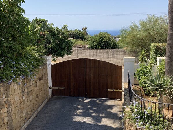 A gated entrance with a wooden gate, a stone wall, and a paved driveway leading to a residential area with greenery and a view of the sea. - Exclusive Duplex Penthouse with panoramic views (Marbella)