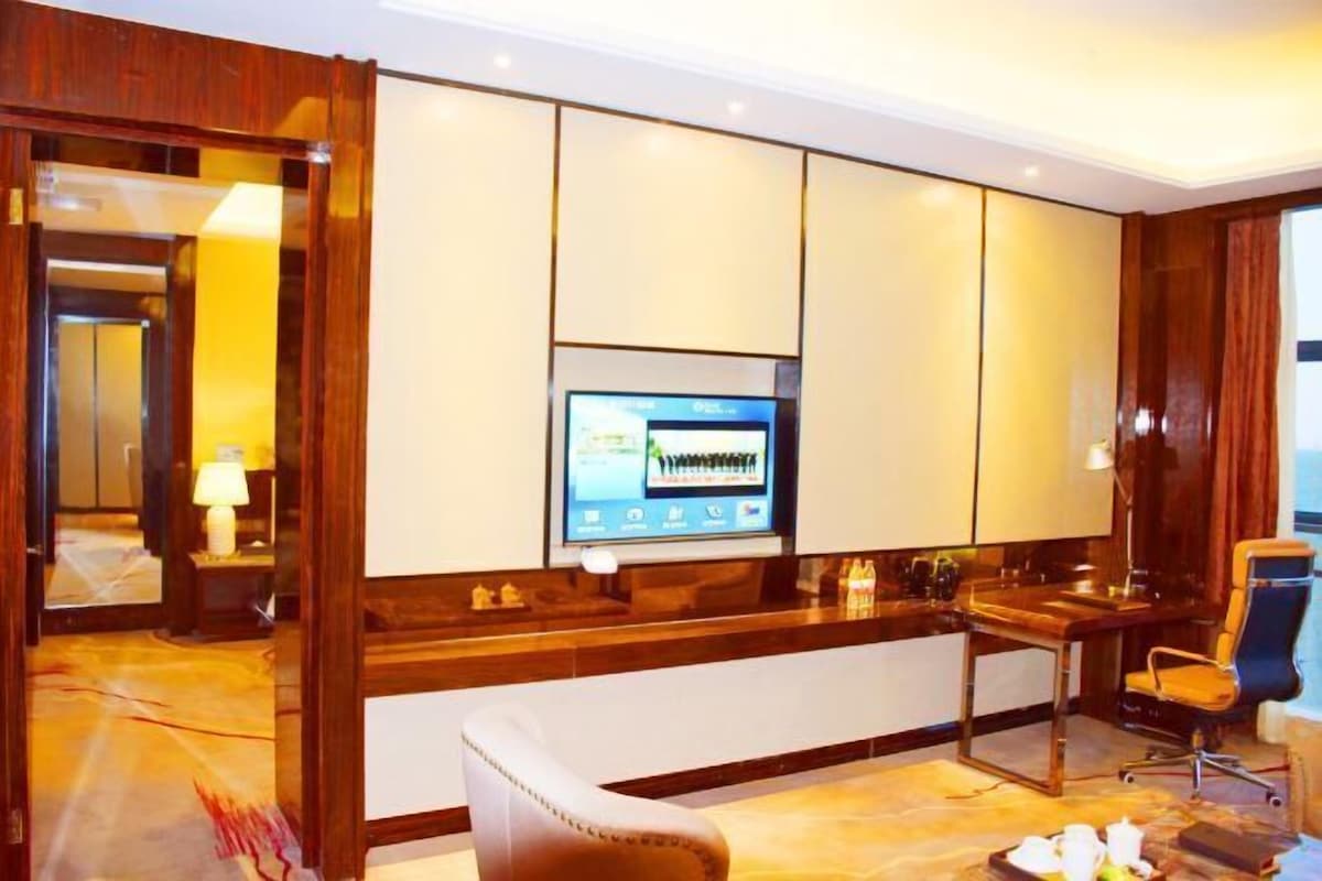 A hotel room with a wooden desk, a television mounted on the wall, a chair, and a small table with a tray of items.
