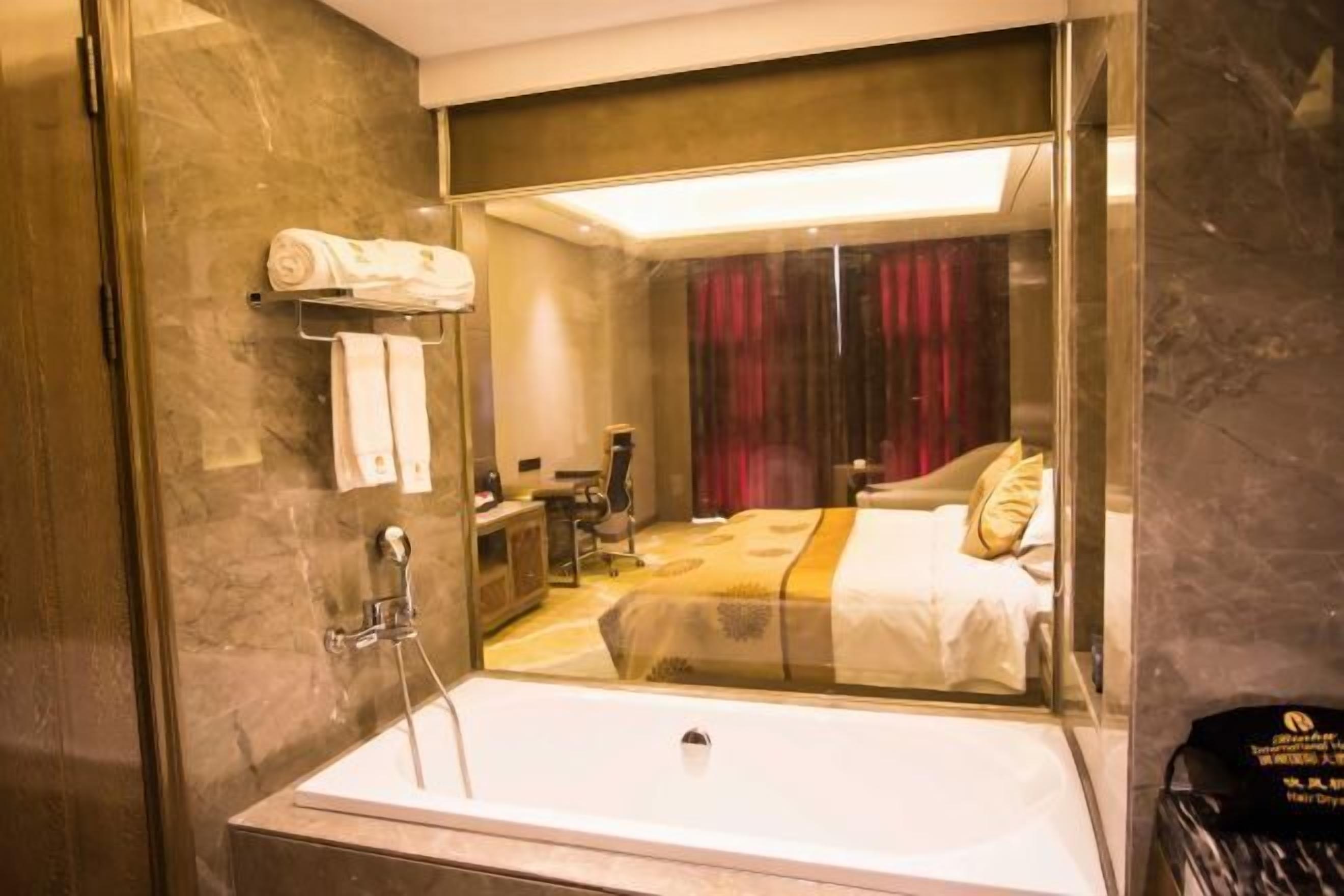 A modern bathroom with a glass-enclosed shower, a white bathtub, and a visible bed with a red curtain in the background.