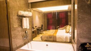 A modern bathroom with a glass-enclosed shower, a white bathtub, and a visible bed with a red curtain in the background.