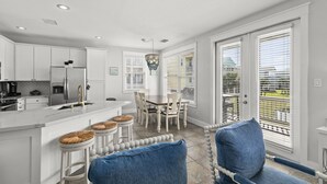 A modern kitchen with white cabinets, a stainless steel refrigerator, and a marble countertop. There are two bar stools with woven seats and a dining table with four chairs. - Beautiful Pointe West Beach House w/Great Sunsets, Beach, Bay Views & Pet Ok (Galveston)