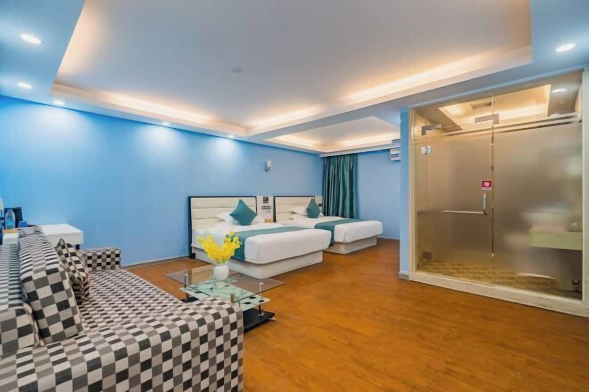 A hotel room with a checkered sofa, a bed with teal bedding, a glass elevator, and a wooden floor.