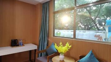 A room with a wooden wall, a window with a view of trees, a small table with a vase of flowers, and a chair with a cushion.