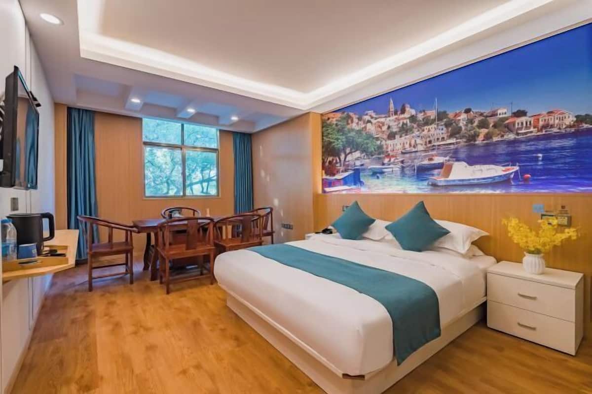A hotel room with a large bed, a desk with chairs, a window with curtains, and a painting of a coastal scene.