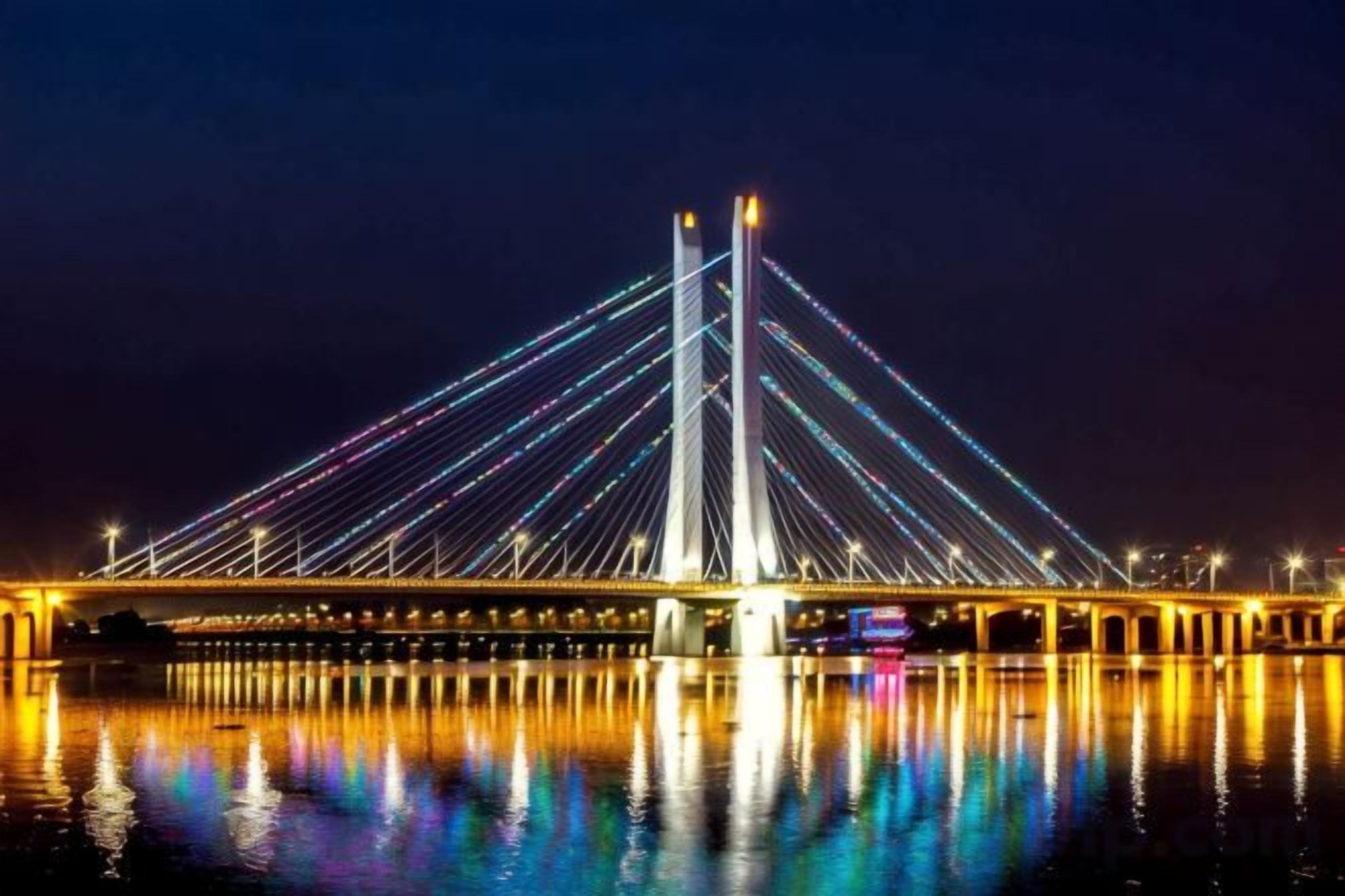 A nighttime view of a cable-stayed bridge with its lights reflecting on the water.
