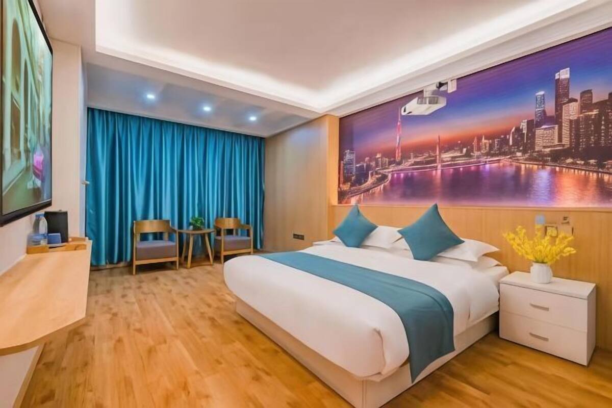 A hotel room with a large bed, a desk, two chairs, a nightstand, and a cityscape mural on the wall.