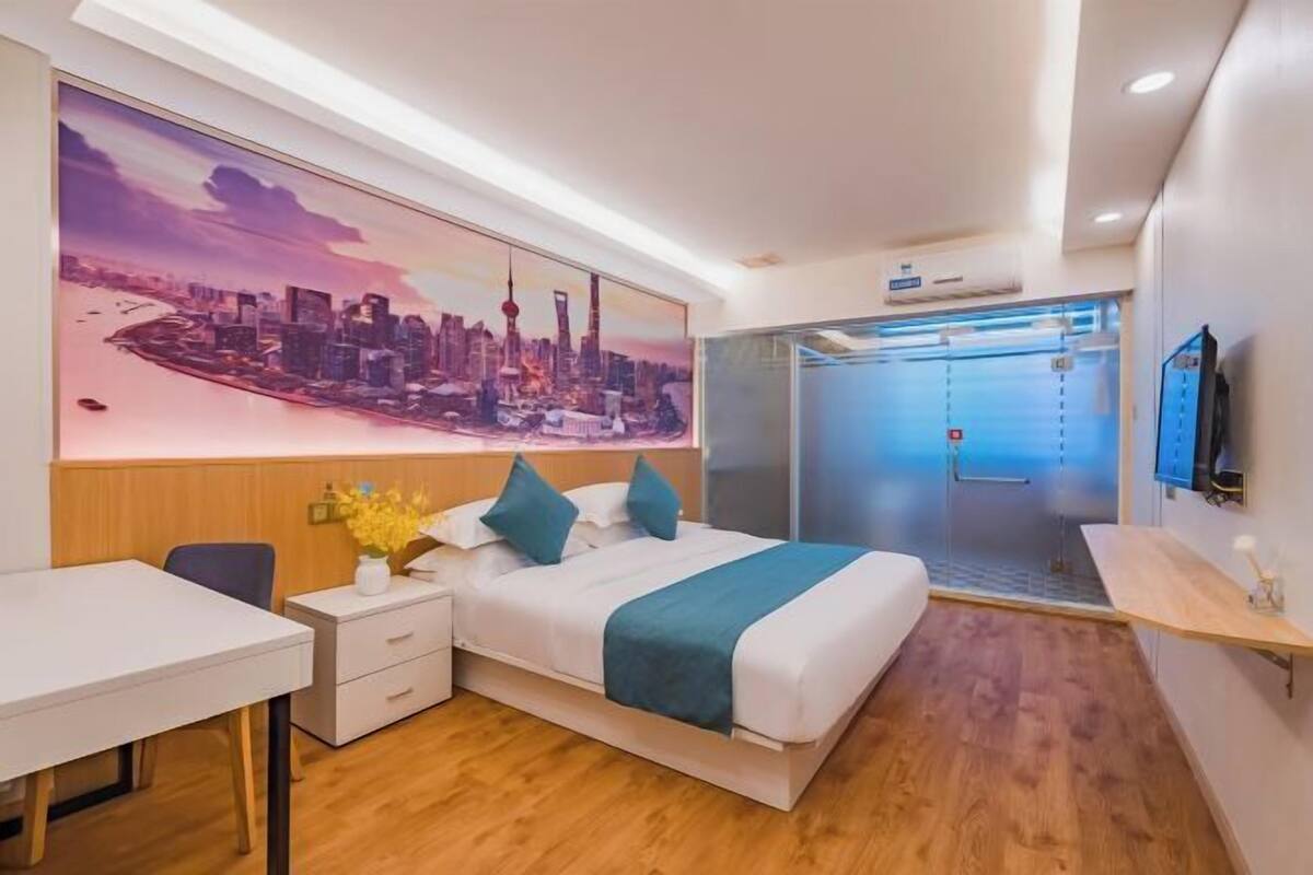 A modern hotel room with a bed, a desk, a chair, a TV, and a large cityscape painting on the wall.