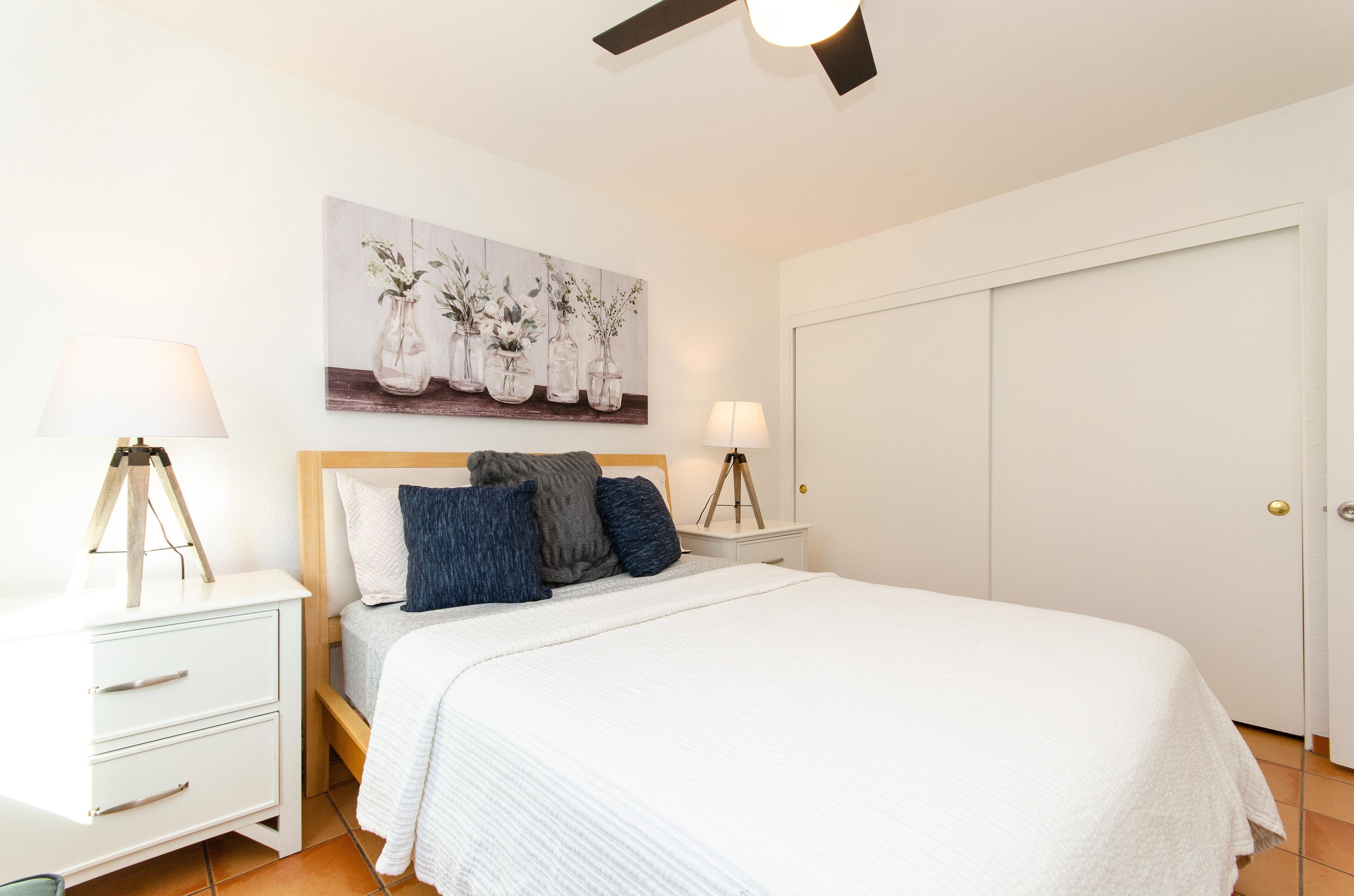 Un dormitorio con una cama, dos mesitas de noche, una lámpara de pared, un ventilador de techo y una puerta.